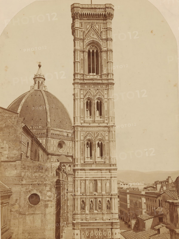 FIRENZE. CAMPANILE DI GIOTTO, Campanile di Giotto and the dome of the Cathedral in Florence, photograph, number: '3018.', Florence, c. 1875 - c. 1900, photographer, Tuscany, paper, pasteboard, albumen print, height 247 mm x width 183 mm, parts of church exterior and annexes: campanile, A sepia-toned photograph shows a tall ornate bell tower with patterned stonework, arched windows, and decorative columns adjacent to a large domed structure with a lantern topped by a small cross. The tower features multiple levels of intricate geometric and floral ornamentation and niches containing sculpted figures. The dome has visible ribs and a tiled surface, with patterned lower walls and round windows. In the foreground there are textured stone walls and rooftops of nearby buildings, and a narrow street lined with multi-story structures receding beside the tower. The sky is empty and pale, occupying the upper background.