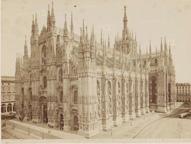 Milan Cathedral, MILAN. LA CATTEDRALE DAL PALAZZO REALE, photograph, number: '3818.', Milan, c. 1875 - c. 1900, photographer, Lombardy, paper, pasteboard, albumen print, height 185 mm x width 248 mm, church (exterior), Dom (Milan), A large Gothic cathedral façade fills the frame with numerous tall pointed spires and pinnacles rising from ornate buttresses and decorative vertical elements; the stone exterior shows many carved statues set into niches, arched doorways with sculpted surrounds, and traceried windows with pointed arches. The foreground shows a paved plaza with steps leading up to the building, a low railing, and a few small groups of people standing or sitting near the entrances. To the right, the cathedral continues with more flying buttresses and a tall central tower topped by a slender spire. Along the left and right edges of the image, multi-story buildings with rows of windows and arcades are visible, and a few street lamps and horse-drawn carriages can be seen near the...