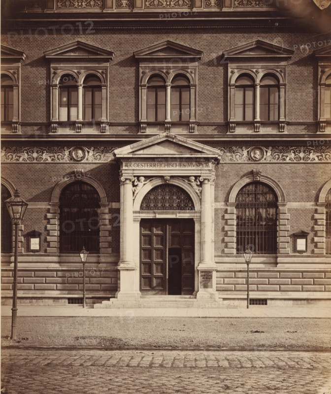 Façade of the Österreichisches Museum für angewandte Kunst in Vienna, Oesterr. Museum for Art and Industry, DETAIL', photograph, number: 'Nr. 64.', Vienna, publisher: Ludwig Niernberger, c. 1875 - c. 1900, Ludwig Niernberger, photographer, publisher, Lower Austria, paper, pasteboard, albumen print, height 233 mm x width 193 mm, column, pillar, architecture, façade (of house or building), A symmetrical stone and brick building façade with a central ornate entrance framed by columns and a triangular pediment, flanked by arched windows with decorative keystones and iron grilles, a frieze of sculpted relief running above the entrance and windows, pairs of tall rectangular windows with rounded tops and paired columns on the upper level, stone steps leading to a large paneled wooden door set within an arched opening with an ornate grille at the top, two cast iron street lamps mounted on poles in front of the building, a paved sidewalk and a cobbled street in the foreground, and decorative ar...