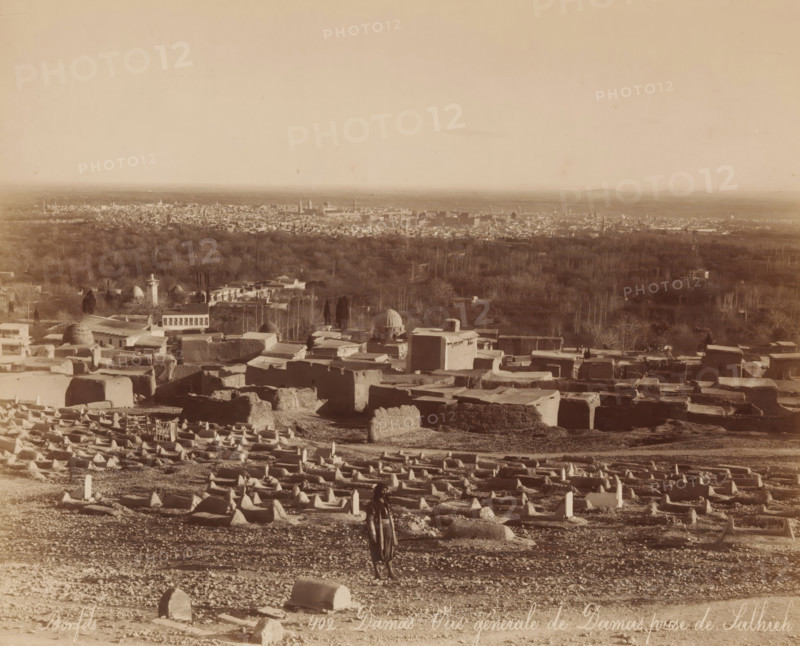 View of Damascus from Salhieh, Damas - Vue générale de Damas prize de Salhieh, photograph, number: '402.', name: 'JAG DE LEUR Représentant Huigenspark, 40b. THE HAGUE.', photographer: Maison Bonfils, Damascus, c. 1875 - c. 1900, Ottoman Empire, paper, pasteboard, albumen print, height 217 mm x width 270 mm, city-view, and landscape with man-made constructions, A wide sepia-toned landscape showing a foreground filled with numerous low rectangular and mound-shaped stone structures arranged in rows on uneven ground, with a solitary person standing among them wearing loose clothing and a head covering, a midground of clustered flat-roofed buildings and domed structures interspersed with trees, and a distant city skyline with many small buildings and taller spires on the horizon beneath a vast pale sky.