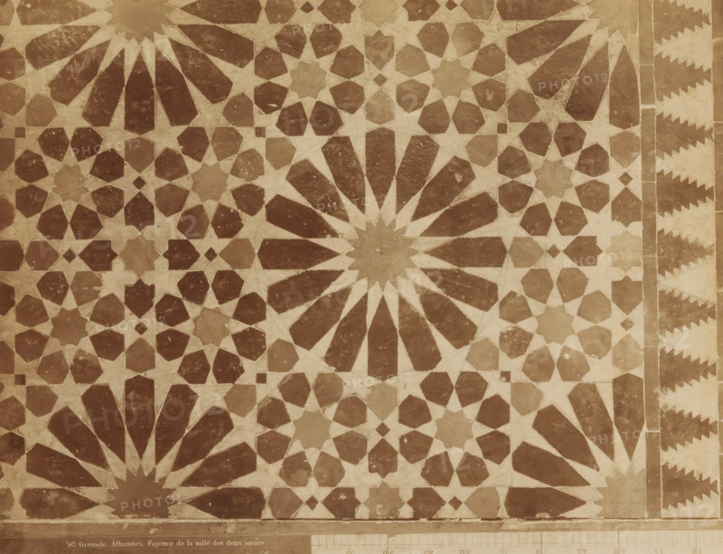 Mosaic of faience in the Hall of the Two Sisters in the Alhambra in Granada, Grenade. Alhambra. Fayence de la salle des deux soeurs, photograph, inscription: 'Syria', number: '587', Alhambra, c. 1875 - c. 1900, photographer, Andalusia, Granada, paper, pasteboard, albumen print, height 217 mm x width 279 mm, ornament, starforms, art (mosaic making), flat decoration, applied arts (Islamic art), A flat surface covered with a repeating geometric mosaic pattern in two contrasting tones, featuring large radial starburst shapes with elongated petal-like segments, interlocking smaller star and polygon motifs, and a narrow decorative border with stepped triangular shapes along one edge, all showing slight surface wear and subtle variations in color and texture.