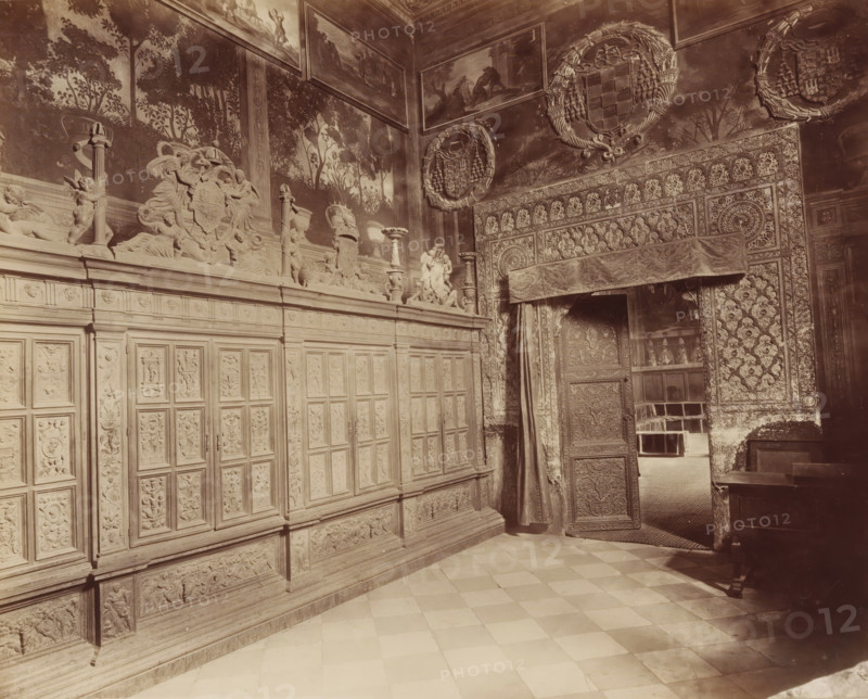 TOLÈDE. LA CATHÉDRALE, Entrance hall to the chapter house of the Cathedral of Toledo, photograph, number: '1396', inscription: 'Spain', Toledo Cathedral, c. 1875 - c. 1900, photographer, Toledo, Castilla-La Mancha, paper, pasteboard, albumen print, height 220 mm x width 277 mm, vestibule, hall, interior of church, painting, applied arts, art (relief, sculpture), A sepia-toned interior view showing an ornately carved wooden wall with rectangular panels and decorative reliefs, a tiled floor in a checkerboard pattern, and an open carved door partially covered by a patterned drape leading into an adjacent room; the upper walls display detailed sculptural friezes with figures, vases, and cherubic forms, painted murals of trees and foliage, framed landscape paintings near the ceiling, and large circular wreath-like medallions with heraldic designs, while the doorway surround and adjacent wall feature dense geometric and floral carved patterns and textiles.
