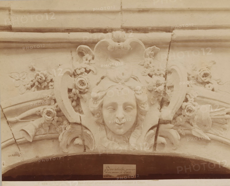 XVIIth century - Bas-relief provenant d'une maison à Caen., Bas-relief with a woman's face, from a house in Caen, photograph, name: 'A. GIRAUDON EDITOR 15. R. BONAPARTE. PARIS', inscription: 'pl 19', number: 'T 424', photographer: Adolphe Giraudon, Paris, c. 1875 - c. 1900, paper, pasteboard, albumen print, height 196 mm x width 241 mm, flowers, ornament, mask, mascaron, sculpture (relief, sculpture), A carved stone relief showing a stylized human face with closed eyes and a slight smile, framed by flowing hair and ornamental scrolls, surrounded by carved flowers and leaves, with curved architectural molding above and beneath and visible cracks and weathering on the surface.