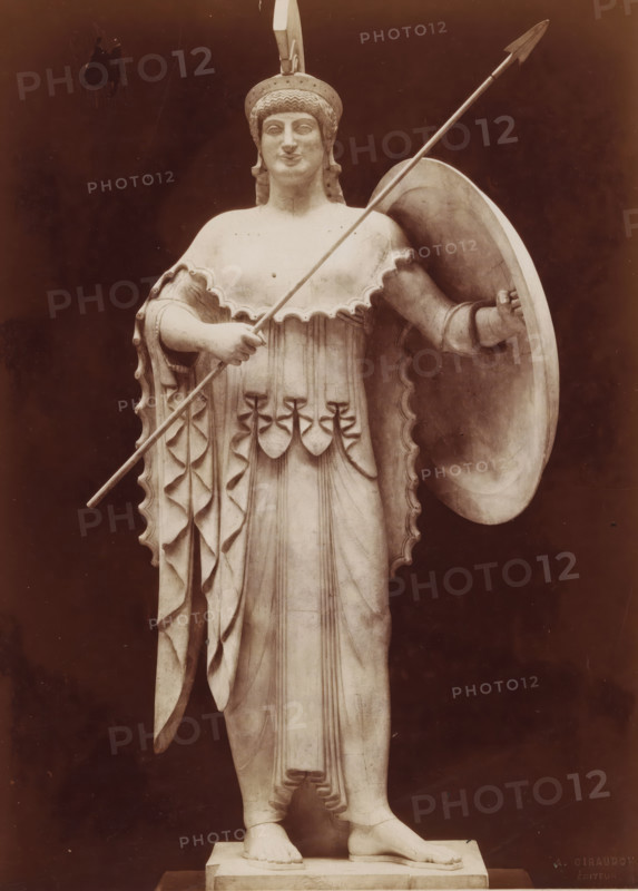 Sculpture of Pallas Athena, from the temple in Aegina, Art Grec. - Athenée provenant du Temple d'Egine Musée de Munich, photograph, name: 'A. GIRAUDON EDITOR 15. R. BONAPARTE. PARIS', inscription: 'pl 4 style k.', number: 'T 10', photographer: Adolphe Giraudon, publisher: Adolphe Giraudon, Paris, c. 1875 - c. 1900, Adolphe Giraudon, photographer, publisher, paper, pasteboard, albumen print, height 258 mm x width 189 mm, sculpture, (story of) Minerva (Pallas, Athena), protective weapons: shield, helved weapons, polearms (for striking, hacking, thrusting): lance, A full-length marble statue of a robed female figure wearing a crested helmet, holding a long spear diagonally across her body and supporting a large round shield at her side, with a scalloped edged cloak draped over her shoulders and detailed folds and decorative elements on the dress, standing barefoot on a rectangular plinth against a dark background.