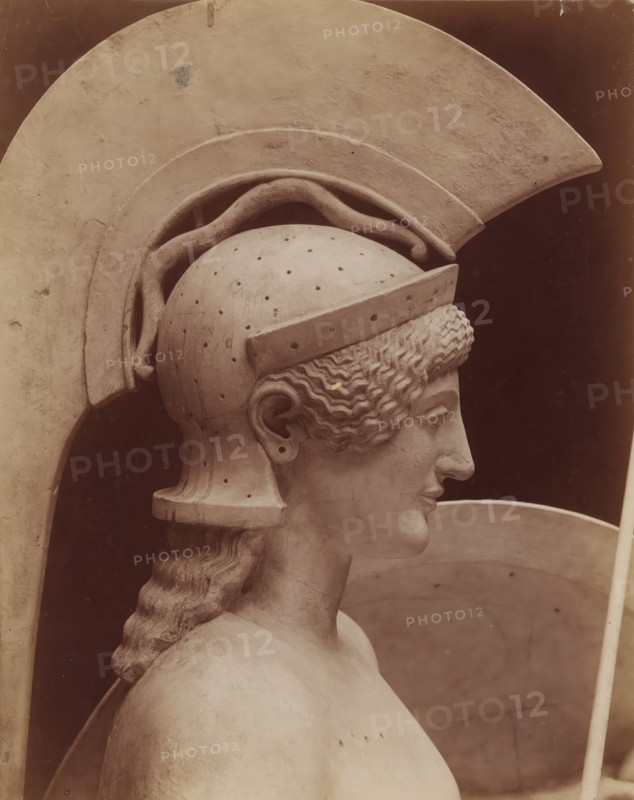 Head of the statue of Pallas Athena, from the pediment of the temple at Egina, Art grec - Tête d'Athene. Fronton du temple d'Egine. Munich, photograph, number: 'T 570 A', inscription: 'pl 4 style k', name: 'A. GIRAUDON EDITOR 15. R. BONAPARTE. PARIS', photographer: Adolphe Giraudon, Paris, c. 1875 - c. 1900, paper, pasteboard, albumen print, height 257 mm x width 199 mm, sculpture, helmet, head (human), (story of) Minerva (Pallas, Athena), A close-up profile of a pale stone sculpture showing a head wearing a large crested helmet with a curved plume, a textured band above the brow, and small perforations across the dome, the face turned slightly forward with a straight nose and closed lips, wavy hair peeking from beneath the helmet and gathered into a long braided lock down the back, a smooth shoulder and upper torso visible, and broad rounded armor or shield elements framing the head against a dark background.