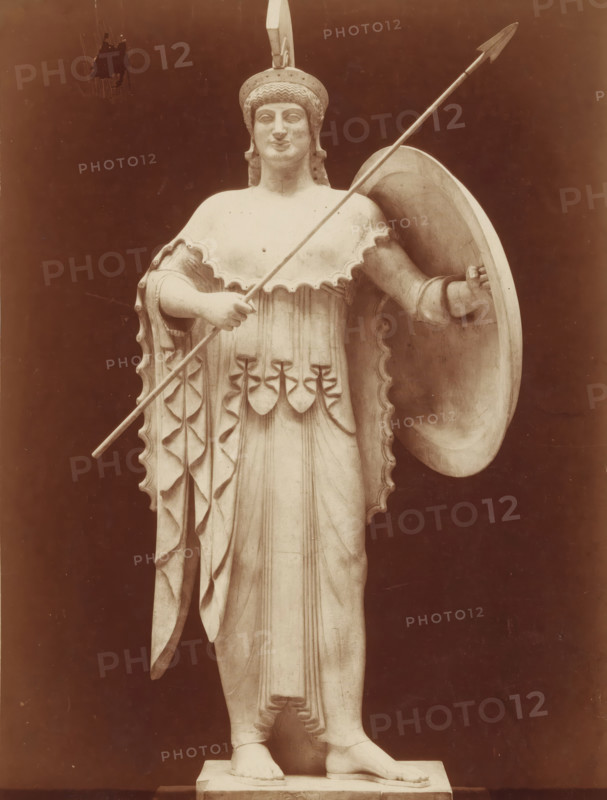 Art Grec Athenée provenant du Temple d'Egine Musée de Munich, Statue of Pallas Athena, from the temple in Egina, photograph, inscription: 'pl 4 style k', number: 'T 570', photographer: Adolphe Giraudon, c. 1875 - c. 1900, Adolphe Giraudon, photographer, paper, pasteboard, albumen print, height 258 mm x width 189 mm, sculpture, (story of) Minerva (Pallas, Athena), protective weapons: shield, helved weapons, polearms (for striking, hacking, thrusting): lance, A full-length marble statue of a robed figure wearing a helmet with a crest, standing on a rectangular plinth against a dark background; the figure holds a long spear diagonally across the body with one hand and a large round shield with the other, the shield resting near the shoulder; the garment features scalloped edges and vertical folds with decorative hanging elements at the waist, and the figure wears sandals with visible toes; the facial expression is calm and the hair is styled beneath the helmet.