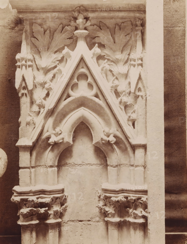 Art Gothique - XIIIe siècle - Pied driot d'une porte. Eglise Saint-Urbain, à Troyes., Part of a doorpost of the Basilica of St. Urbanus in Troyes, photograph, name: 'A. GIRAUDON EDITOR 15. R. BONAPARTE. PARIS', number: 'T 1224', collector's mark: 'Diary and Art Craft School for Girls Amsterdam', collector's mark: 'INSTITUTE FOR ART-INDIVIDUAL EDUCATION GABRIËL METSUSTRAAT No. 16, AMSTERDAM', photographer: Adolphe Giraudon, publisher: Adolphe Giraudon, Paris, c. 1875 - c. 1900, Adolphe Giraudon, photographer, publisher, paper, pasteboard, albumen print, height 258 mm x width 203 mm, foliage, tendrils, branches, ornament, stile, post, doorjamb, column, pillar, architecture (Gothic art), A close-up of a carved stone architectural fragment showing a pointed arch framed by layered moldings and ornamental foliage; the arch has a trefoil motif set within a triangular gable decorated with small bud-like finials and stylized leaf carvings extending outward; beneath the arch are clustered column...