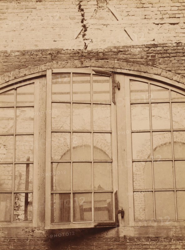 Detail of a window section at the Binnenhof during the restoration, photograph, annotation: 'Courtyard between the Knight's Hall and the Roll Room, photo taken there', annotation: 'Count's Halls, Restoration of the courtyard between the Knight's Hall and the Roll Room, April 1899, The Hague', Binnenhof, 1899, photographer, The Hague, Attributed to, paper, pasteboard, albumen print, height 224 mm x width 167 mm, closed window, A large arched multi-pane window set into a brick wall, with wooden frames and muntins dividing the glass into rectangular sections, some panes showing reflections and textured surfaces behind them, a vertical wooden post between the window sections, metal hinges and a latch attached to the frames, weathered brickwork above and below the window with visible mortar lines and a horizontal stone lintel, areas of chipped and worn paint and surface irregularities on both wood and brick.
