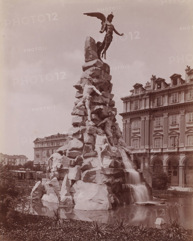 Piazza dello Statuto - Momumento del Frejus, View of the Momumento del Frejus at the Piazza Statuto in Turin, Italy, Torino, photograph, number: '8106a).', inscription: 'Brogi (t), MB', photographer: attributed to Carlo Brogi, Turin, publisher: attributed to Edizioni Brogi, Florence, after sculpture by anonymous, 1881 - 1900, after sculpture by, Tuscany, Piemonte, Attributed to, pasteboard, paper, albumen print, albumendruk, height 445 mm x width 318 mm, piece of sculpture, reproduction of a piece or sculpture, monument, statue, ornamental fountain, Piedmont, Piazza Statuto, A tall sculpted rock formation rises from a pool with water cascading down its side, populated by several human figures carved in classical style and a winged figure poised above the peak; the rocky structure and figures occupy the left and center of the image while a multi-story building with rows of windows, ornate cornices, and dormer windows fills the right background; plants and grasses line the pond in the fo...