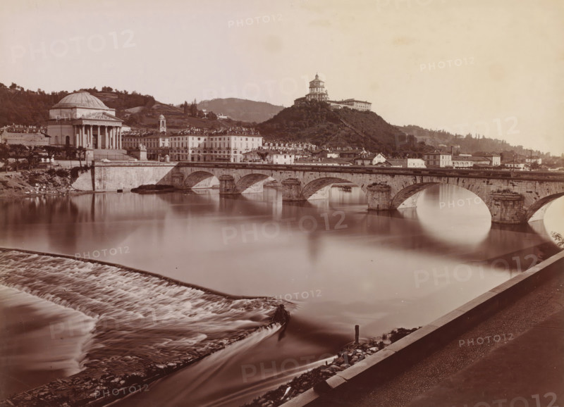La gran Madre di Dio, View of the Po and the Gran Madre di Dio in Turin, Italy, Torino, photograph, number: '3705.', number: '88', photographer: Giacomo Brogi (possibly), Turin, publisher: attributed to Edizioni Brogi, Florence, 1864 - 1881, Tuscany, Piedmont, Attributed to, pasteboard, paper, albumen print, height 317 mm x width 455 mm, landscape with bridge, viaduct or aqueduct, river, temple, shrine, non-Christian religions, Piemonte, A wide riverscape showing a calm reflective body of water crossed by a stone arched bridge with multiple semicircular openings and sturdy piers, a stepped weir creating cascading water in the foreground, and a riverbank promenade with a paved walkway. On the far bank a classical domed building with a columned portico stands beside a row of multi-story buildings with uniform facades and a smaller bell tower, while a tree-covered hill rises behind them topped by a large hilltop structure with a tiered profile. Hills and scattered trees extend into the ba...