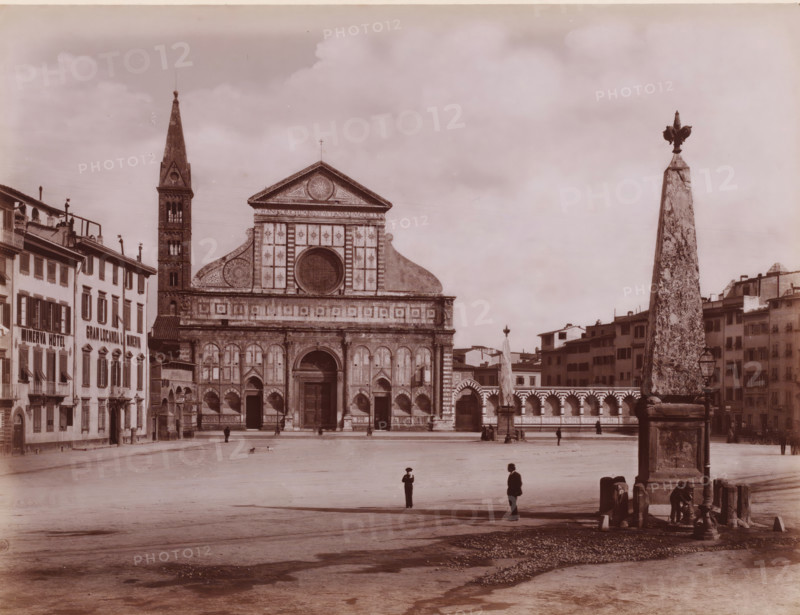 Facciata della Chiesa di Santa Maria Novella, compiuta da LB Alberti?, View of the Santa Maria Novella in Florence, Italy, Firenze, photograph, number: '3027.', number: '111', photographer: Giacomo Brogi (possibly), Florence, publisher: attributed to Edizioni Brogi, after sculpture by Giambologna, 1864 - 1881, Giambologna, after sculpture by, Tuscany, Attributed to, pasteboard, paper, albumen print, height 318 mm x width 445 mm, church (exterior), Santa Maria Novella, A wide plaza with a large ornate stone church facade centered in the background, featuring a tall narrow bell tower to its left and decorative arches and a circular window above the main entrance, flanked by rows of multi-story buildings with shuttered windows on the left and right; an obelisk with a sculpted top stands prominently on the right foreground near a small fountain where a person is leaning, and a few scattered people and small animals are visible across the open paved square beneath a cloudy sky.
