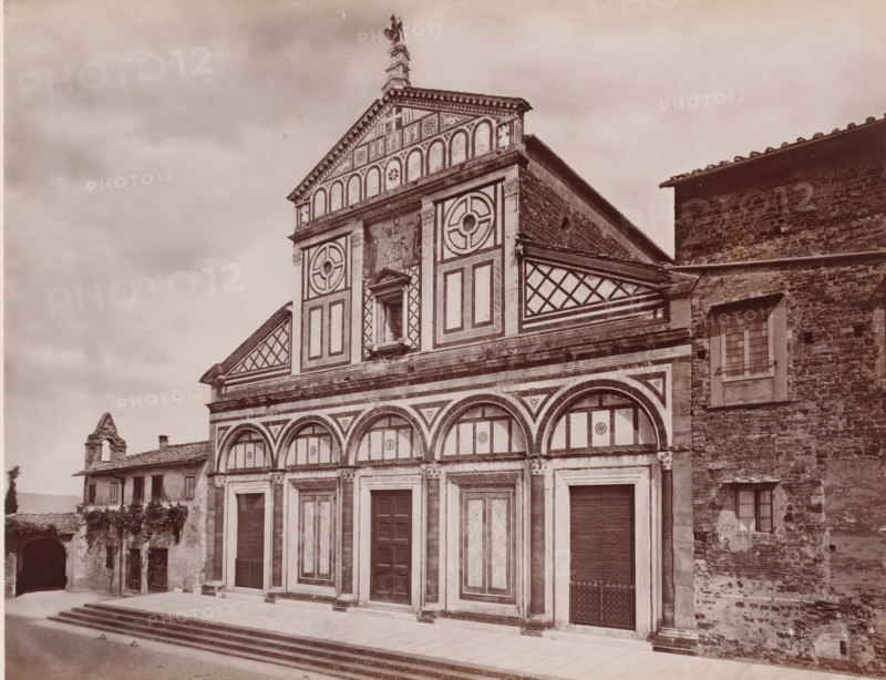 Chiesa di S. Miniato di stile greco romanzo, anteriore al mille, Façade of the San Miniato al Monte in Florence, Italy, Firenze, photograph, number: '113', number: '3031.', photographer: Giacomo Brogi (possibly), Florence, publisher: attributed to Edizioni Brogi, 1864 - 1881, Tuscany, Attributed to, pasteboard, paper, albumen print, height 318 mm x width 445 mm, church (exterior), S. Miniato, A large stone building façade with geometric and arched decorative panels, tall doorways set into framed rectangular bays, and a tiered upper section featuring circular motifs and patterned panels; a central niche with a sculpted figure flanked by painted or carved figures sits above the middle doorway, and a triangular pediment tops the composition with ornamental detailing and a statue at its peak. To the right, an adjoining stone wall with small windows and a tiled roof contrasts with the patterned façade, while to the left a smaller building with shuttered windows, a tiled roof, and an arched ...