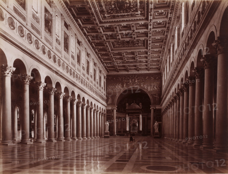 Interior of St. Paul Outside the Walls in Rome, Italy, Interno della Basilica di S. Paolo fuori le mura, Roma, photograph, inscription: 'Roma, S. Paolo', number: '8018.', number: '56.', photographer: Carlo Brogi, Florence, publisher: Edizioni Brogi, 1881 - 1900, Tuscany, pasteboard, paper, albumen print, height 317 mm x width 445 mm, interior of church, Rome, Saint Paul outside the Walls, A wide interior view of a large hall with a reflective patterned floor leading toward an ornate apse, flanked on both sides by tall rows of smooth columns topped with decorative capitals supporting arcades; above the arcades the walls display framed oval medallions and rectangular panels interspersed with arched windows, and the coffered ceiling is richly decorated with geometric and floral reliefs; at the far end an archway with a large painted image and sculptural figures frames an elevated altar area with ornate furnishings, and two freestanding statues stand near the entrance to that area.