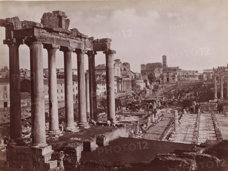 Fôro Romano, Roman Forum with Temple of Saturn in Rome, Italy, Roma, photograph, number: '103.', number: '3643.', photographer: Giacomo Brogi (possibly), Rome, publisher: attributed to Edizioni Brogi, Florence, 1864 - 1881, Tuscany, Attributed to, pasteboard, paper, albumen print, height 317 mm x width 445 mm, ruin of a building, architecture, landscape with ruins, Roman Forum, Temple of Saturn, A wide sepia-toned panorama of ancient ruins dominated on the left by a row of tall classical columns with carved capitals and a partially intact entablature, standing on heavy bases above worn stone foundations, with scattered broken masonry and rubble at their feet; behind and between the columns are low buildings with tiled roofs and simple facades, and further back a domed structure and a taller multi-tiered tower rise above other classical and baroque façades; the central and right areas show an expanse of archaeological remains including fragmented walls, paved pathways, rows of short rui...