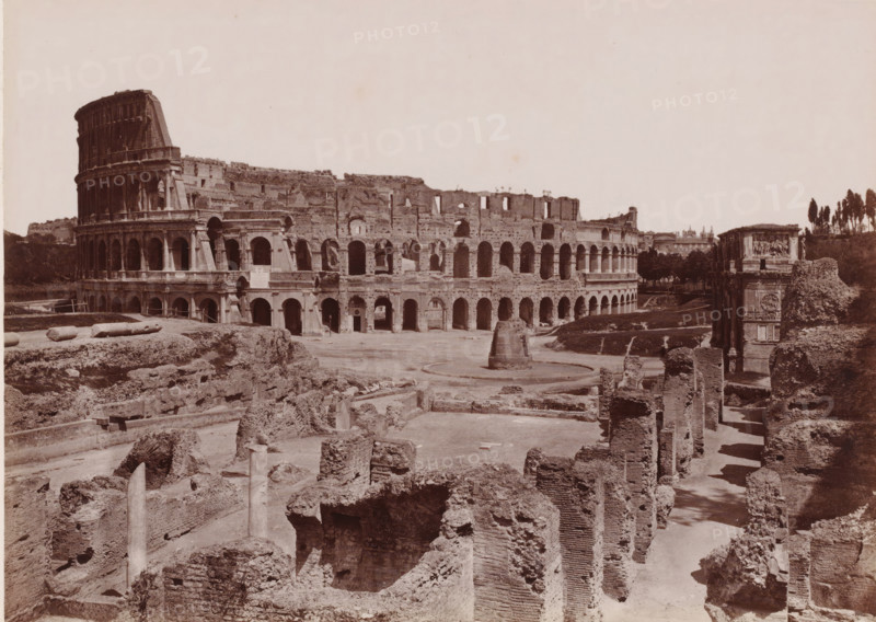 View of the Colosseum in Rome, Italy, Colosseo, Roma, photograph, number: '4222.', number: '104.', photographer: Giacomo Brogi (possibly), Rome, publisher: attributed to Edizioni Brogi, Florence, 1864 - 1881, Tuscany, Attributed to, pasteboard, paper, albumen print, albumendruk, height 317 mm x width 445 mm, ruin of a building, architecture, landscape with ruins, amphitheatre, Colosseum, A wide sepia photograph shows an ancient stone amphitheater with multiple tiers of arched openings and a partially ruined outer wall, foregrounded by excavated ruins of brick and stone foundations and scattered columns. The amphitheater curves across the scene with a rounded, taller section on the left and a lower, arcaded stretch toward the right. In front of the amphitheater there is an open dirt area with a circular raised stone feature and a freestanding cylindrical structure. To the right of the amphitheater stands a decorated stone arch with relief panels, flanked by more rubble and vegetation. T...