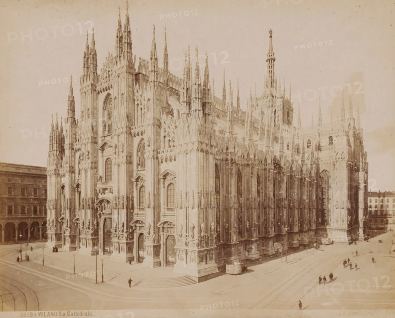 View of the Cathedral of Milan, Italy, La Cattedrale, Milano, photograph, number: '3818a.', collector's mark: 'VOSMAER', Milan, publisher: Giacomo Brogi, Florence, 1856 - 1881, Giacomo Brogi, photographer, publisher, Tuscany, Lombardy, pasteboard, paper, albumen print, height 314 mm x width 420 mm, church (exterior), Cathedral (Milan), A large ornate cathedral façade fills most of the image, showing tall pointed spires, intricate stone carvings, arched windows, and numerous sculpted figures and decorative elements on multiple levels; broad steps lead up to several arched entrances at street level, where small groups of people and a few lone figures are scattered along the sidewalks and near lampposts; a tram or streetcar is visible beside the building and tracks run along the adjacent streets, with clusters of pedestrians walking across open paved areas; to the left and right low multi-story buildings frame the scene, and the overall image has a pale, sepia-toned appearance.