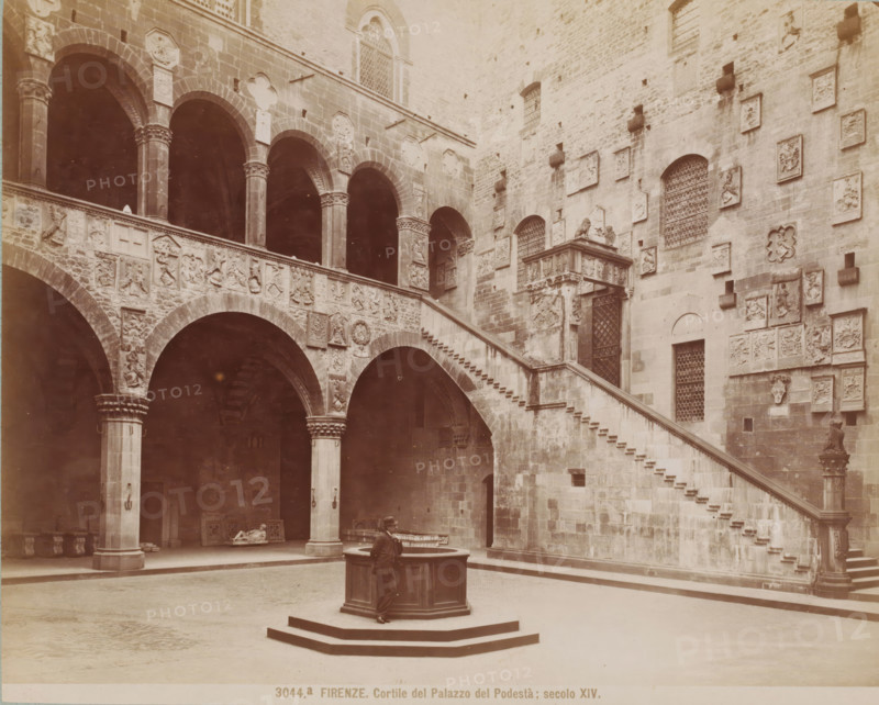 Courtyard of the Palazzo del Podestà in Florence, Italy, Cortile del Palazzo del Podestà, secolo - 1881, Giacomo Brogi, photographer, Florence, Tuscany, Attributed to, pasteboard, paper, albumen print, height 316 mm x width 422 mm, courtyard, palace, Palazzo del Podestà (Florence), A stone courtyard with a central octagonal well on a raised platform and a man leaning on its edge, a wide stair with stone steps ascending along a wall decorated with numerous carved plaques and reliefs, arched openings supported by columns with decorated capitals, a second level balustrade carved with heraldic and figurative reliefs, grated windows set into the masonry, sculptural elements and decorative panels mounted on the walls, dark recessed archways beneath the arcade, and scattered stone artifacts visible in the shaded areas.