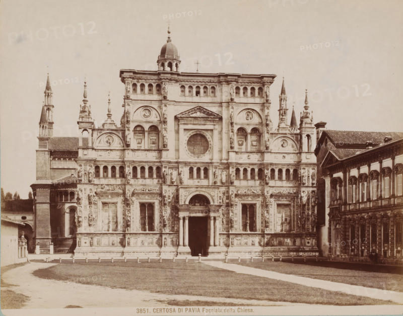 Facciata della Chiesa, Façade of the Certosa di Pavia, Italy, Certosa di Pavia, photograph, collector's mark: 'VOSMAER', number: '3851.', Pavia (city), publisher: attributed to Giacomo Brogi, 1856 - 1881, Giacomo Brogi, photographer, publisher, Lombardy, Attributed to, pasteboard, paper, albumen print, height 315 mm x width 421 mm, abbey, monastery, convent, Roman Catholic church, Pavia (province), A wide frontal view of an ornate stone church facade with multiple levels of arches, columns, statues, and decorative reliefs, flanked by tall spires and smaller towers, a central entrance with columns beneath a pediment and a large circular window above, a domed cupola near the top, adjacent buildings with tiled roofs on either side, a grassy foreground with a curved path leading toward the entrance, and a clear pale sky above.