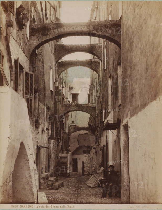Street view with some people in San Remo, Italy, Vicolo del Giuoco della Palla, Sanremo, photograph, number: '8091.', collector's mark: 'VOSMAER', inscription: 'doubl', photographer: Carlo Brogi (possibly), San Remo, publisher: Edizioni Brogi, Florence, 1881 - 1900, Carlo Brogi, Edizioni Brogi, photographer, publisher, Tuscany, pasteboard, paper, albumen print, hand coloring, height 421 mm x width 316 mm, public road, peninsula, A narrow cobblestone alley flanked by tall, weathered buildings with shuttered windows and rough plastered walls, crossed overhead by a series of worn stone arches creating a tunnel-like perspective, with hanging laundry and small balconies visible along the upper levels, a street lamp suspended from one wall, stone steps and low ledges lining the sides, and several people present including two figures looking down from a high window and a seated woman in a long dress with another person nearby, plus a dog standing partway down the alley.