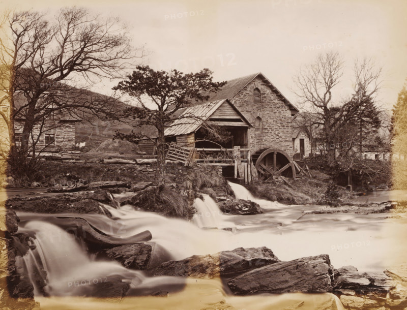 Watermill at the Dochart in Killin, Mill on the Docharts, Killin, album page, photograph, This page is part of an album., number: '746, JV', c. 1875 - c. 1885, photographer, Stirling, paper, albumen print, height 298 mm x width 236 mm, watermill, A stone building with a pitched roof sits beside a flowing stream, a large wooden water wheel attached to its side and a wooden shelter extending from the structure; a tree with bare branches stands in front of the building and another tree with denser foliage is nearer the center, while fallen logs, wooden fences, and rough stone walls are scattered around the yard; cascading water flows over rocks in the foreground forming small waterfalls, and additional leafless trees and shrubs frame the scene on either side under a pale sky.