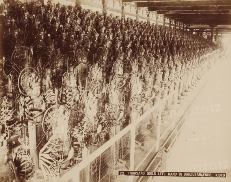 22. Tnousand Idols left hand in Sanjiusangendo.- Kyoto, Wooden statues of Kannon in the Sanjusangendo Temple, Kyoto, photograph, number: '9.', c. 1895 - c. 1915, photographer, Kinki, paper, pasteboard, albumen print, height 210 mm x width 269 mm, height 243 mm x width 329 mm, A sepia-toned photograph showing a long row of similar ornate statues arranged closely together on a raised platform behind a wire mesh railing, each statue featuring detailed headdresses, multiple arms, and circular halo-like structures, with the figures extending into the distance under a ceiling supported by beams and posts, and bright overexposed light on the far right side obscuring details.