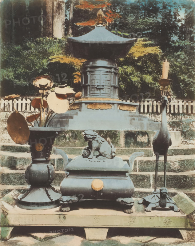 Tomb of Ieyasu, Nikko, photograph, annotation: '38. Tomb.', c. 1895 - c. 1915, photographer, Kanto, paper, pasteboard, salted paper print, Salt print, colored., height 264 mm x width 208 mm, height 329 mm x width 243 mm, A stone and metal shrine setup sits on tiered stone steps with a large ornate metal canopy structure centered toward the top, flanked by dense foliage and trees in the background; in front of the canopy a rectangular pedestal features decorative panels and a rounded top section, and on the lower platform a sculpted guardian lion rests atop a boxy incense burner with curved handles and a round ornament on its front; to the left a tall vase holds dried lotus pods and large round leaves, and to the right a tall, slender metal stand supports a slender candle above a turtle-shaped base; the scene shows weathered surfaces, patinaed metal, mossy stone, and a muted palette of greens, browns, and faded blue-gray tones.