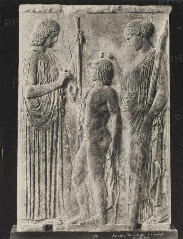 Demeter, Persephone and the child Triptolemos, Déméter, Perséphone & l'enfant Triptolème, photograph, number: '45.', Greece, c. 1895 - c. 1915, photographer, pasteboard, paper, collotype, photoprint, height 280 mm x width 218 mm, height 328 mm x width 239 mm, A carved stone relief showing three figures in profile standing close together against a rectangular panel, with two taller robed figures flanking a smaller nude figure in the center; the taller figures wear long draped garments with detailed vertical folds and hold staffs, one with wavy shoulder-length hair and the other with hair pulled back, while the central figure has short curly hair and a slender youthful body; the taller figures reach toward the central figure with one hand each, their garments showing layered folds and textured fabric, and the surface of the stone exhibits weathered texture and varying tones.