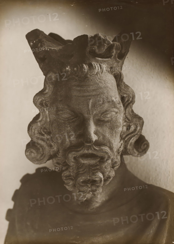 Sculpture in the cloister of Bamberg Cathedral, Cloister, Head, album page, photograph, This photo is part of an album., number: '148.', Bamberg, 1900 - c. 1920, photographer, Bavaria, paper, baryta paper, gelatin silver print, height 225 mm x width 164 mm, interior of church, Bamberg Cathedral, A close view of a weathered stone bust showing an aged face with closed eyes, a pronounced nose, curled hair framing the forehead and temples, a full curled beard with layered ringlets, and a textured crown-like headdress at the top, all rendered in monochrome tones against a softly lit plain background.