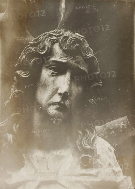 Detail of the Riemenschneideraltar in Bamberg Cathedral, Domschatz, hl. Sebastian (Riemenschneiderschule) (Kopf), album page, photograph, This photo is part of an album., inscription: 'Kunsthist. Seminar, Marburg (Lahn), Photogr. Abteilung, Vervielfältigung, untersagt.', inscription: 'Bamberg, 166', Bamberg, 1900 - c. 1920, photographer, Bavaria, paper, baryta paper, gelatin silver print, developing gelatin silver print, height 236 mm x width 176 mm, interior of church, Bamberg Cathedral, A close-up of a weathered stone or metal sculpture showing a solemn male face with long wavy hair and a small beard, detailed curls framing the forehead and sides, deep-set eyes with heavy lids and a furrowed brow, a pronounced nose and downturned mouth, textured surface with visible patina and wear, parts of the shoulders and upper chest visible with draped garment and carved decorative curls, and a dark, grainy background.