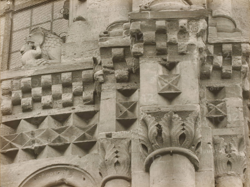 East Church, 1. Frieze 4., View of the sculpture of a frieze of Bamberg Cathedral, album page, photograph, This photo is part of an album., number: '6.', Bamberg, 1900 - c. 1920, photographer, Bavaria, paper, baryta paper, gelatin silver print, developed gelatin silver print, height 165 mm x width 219 mm, church (exterior), Bamberg Cathedral, A close-up view of a weathered stone facade showing carved geometric patterns and architectural details in a monochrome tone, including rows of protruding square blocks, recessed triangular indentations, diamond-shaped reliefs, rounded column segments, and decorative foliage-like carvings, with a sculpted figure of a lion partially visible near a grated window at the top left.