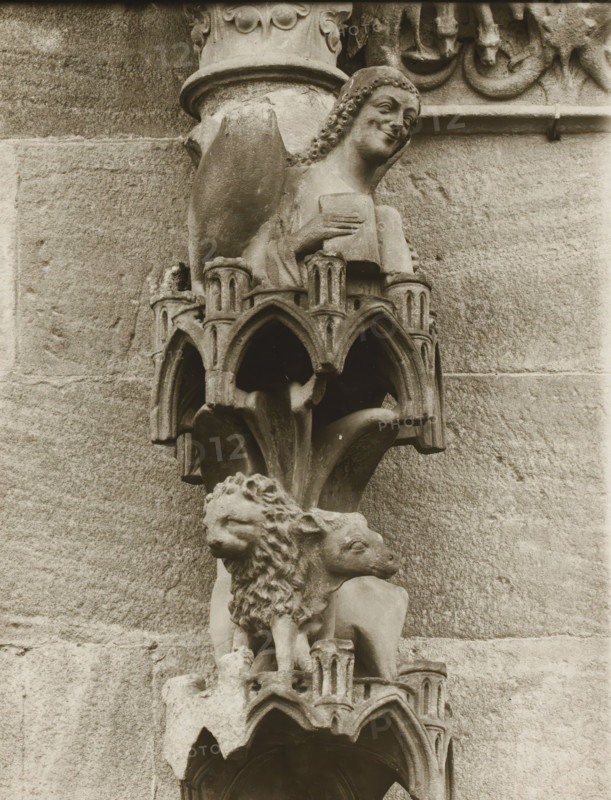 Adamspforte, Engel, Löwe, Ochse vN, Detail of the Fürstenportal in Bamberg Cathedral, album page, photograph, This photo is part of an album., inscription: 'Kunsthist. Seminar, Marburg (Lahn), Photogr. Abteilung, Vervielfältigung, untersagt.', number: '62', Bamberg, 1900 - c. 1920, photographer, Bavaria, paper, baryta paper, gelatin silver print, developing gelatin silver print, height 238 mm x width 181 mm, church (exterior), Bamberg Cathedral, A carved stone figure perched on an ornate bracket against a masonry wall, showing a smiling human face with curled hair and a hand holding a rectangular object, above a detailed canopy of pointed arches and small columns, with a sculpted lion head and an animal head partially visible below, all rendered in weathered stone tones and set against the textured stone blocks of the building.