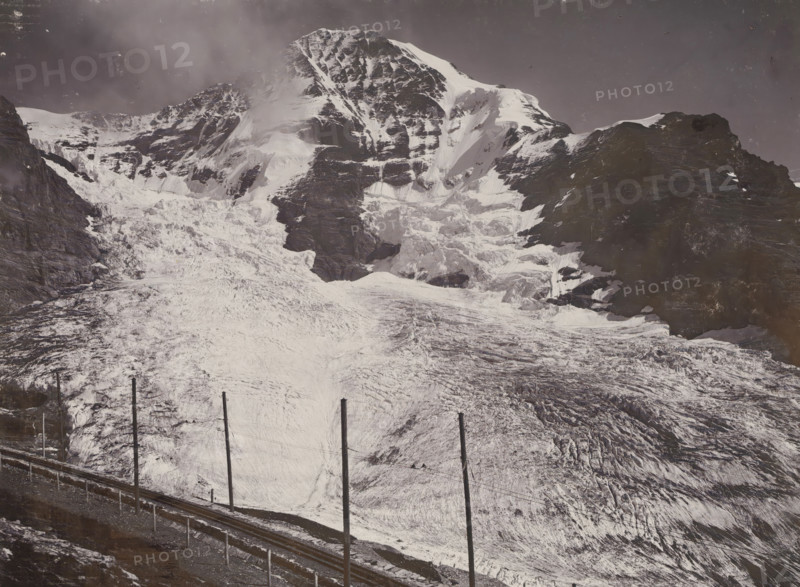 Jungfraubahn und Mönch, View of the Mönch with the Jungfraubahn railway, photograph, This photo is part of a cover., inscription: 'Gebr. Wehrli. Kilchberg-Zurich. 3890. Jungfraubahn und Mönch.', Jungfraujoch, publisher: Gebrüder Wehrli, Kilchberg, 1912 - 1920, photographer, Alpen, Bernese Alps, paper, pasteboard, baryta paper, daylight collodion silver print, height 164 mm x width 218 mm, railway, train, mountain-top, peak, Alps, Mönch, A wide snowy mountain scene fills the image, showing a steep, rugged peak with layered rock faces and snow fields, a large glacier flowing down the central slope with crevasses and textured ice, darker rocky ridges flanking the snowy areas, and a railway track with tall vertical poles running along the lower foreground beside a narrow ledge.