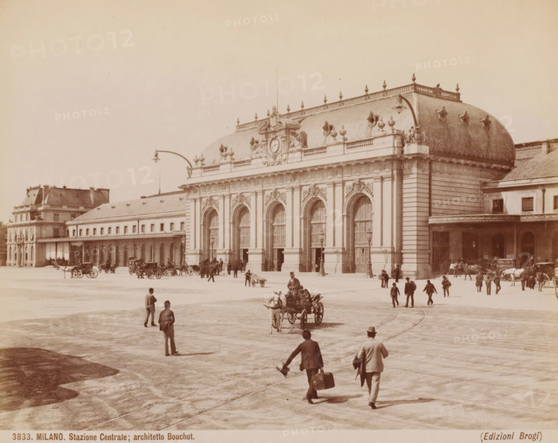 View of Milan station, Italy, Stazione Centrale, architetto Bouchot, Milano, photograph, inscription: '(Diepr. nakomen)', number: '3833.', photographer: Giacomo Brogi (possibly), Milan, publisher: Edizioni Brogi, Florence, after design by Louis-Jules Bouchot, 1864 - 1881, Tuscany, Lombardy, pasteboard, paper, albumen print, height 251 mm x width 331 mm, station, shelter (railway, tramway), exterior, representation of a building, Milano Centrale Station, A wide plaza in front of a large ornate stone building with tall arched doorways and decorative sculpted details along its façade, a curved mansard roof with ornamental urns and a central clock-like round relief, many people walking or standing across the open paved area, several horse-drawn carriages and horses near the building, a few lampposts mounted on curved arms projecting over the plaza, smaller adjacent buildings flanking the main structure, and distant figures and carts near the building edges.