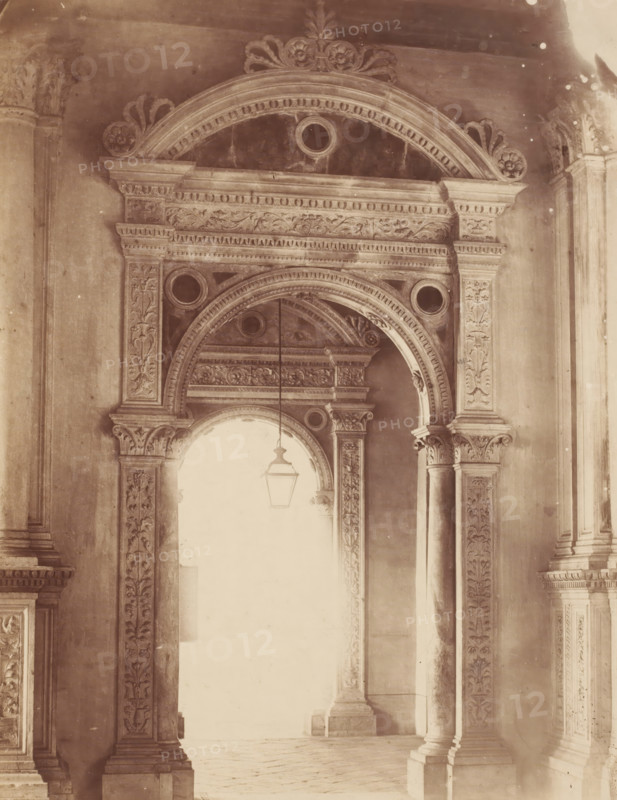 Gate with lantern in Venice, Italy, photograph, inscription: 'RSKN 1', collector's mark: 'RICH SCHOOL FOR ART HANDBOOK, GROUP', inscription: 'Venice', inscription: 'I', Venice, 1851 - 1900, photographer, pasteboard, paper, albumen print, height 479 mm x width 318 mm, gallery (spacious corridor), A sepia-toned photograph of an ornate architectural interior showing carved stone arches and columns with floral and scrollwork decoration, a large rounded archway framed by detailed molding and circular recessed ornaments, a hanging lantern suspended in the open arch, fluted columns with decorative capitals and carved vertical panels, and a bright lighted space visible through the arch contrasting with the darker carved surfaces.