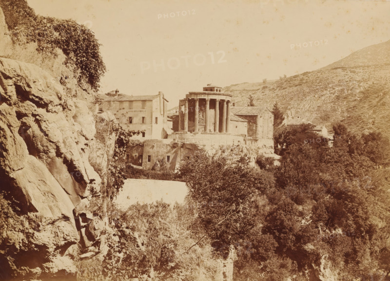 View of the Temple of Vesta at Tivoli, photograph, photographer: attributed to Giorgio Sommer, c. 1860 - c. 1875, Attributed to, height 270 mm x width 369 mm, height 475 mm x width 615 mm, A sepia-toned scene showing a rocky cliff face on the left with vegetation clinging to it, a cluster of buildings perched on a ledge toward the center including a round classical temple with tall columns and a tiled-roof structure adjacent to it, trees and shrubbery filling the foreground and lower right, a steep hillside rising in the background on the right, and uneven rocky terrain and foliage across the lower part of the image.