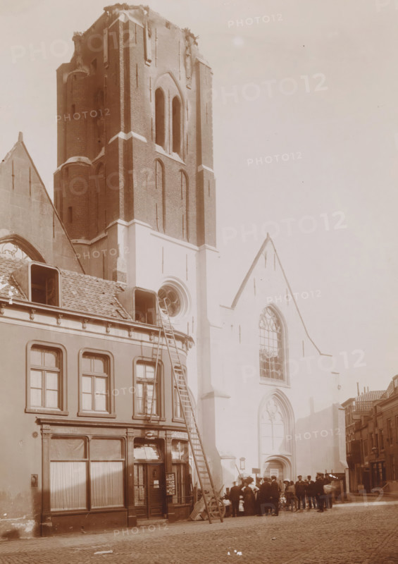 View of the Reformed Church in Vlissingen after the fire in 1911, photograph, annotation: 'JB van Bree, 713d, 2554', annotation: 'NH church in Vlissingen after the fire of September 6, 1911', Flushing, 1911, photographer, Attributed to, paper, pasteboard, daylight collodion silver print, height 211 mm x width 168 mm, church (exterior), street (city(-scape) with figures, staffage), A tall brick church tower with arched openings rises beside a lighter stone facade with large pointed arched windows; in front of the building a row of narrow townhouses with tiled roofs and dormer windows lines the street, one with a tall ladder leaning against it; a group of people stand near the church entrance, some wearing hats and a bicycle nearby; cobbled street in the foreground and additional buildings receding along the right side.