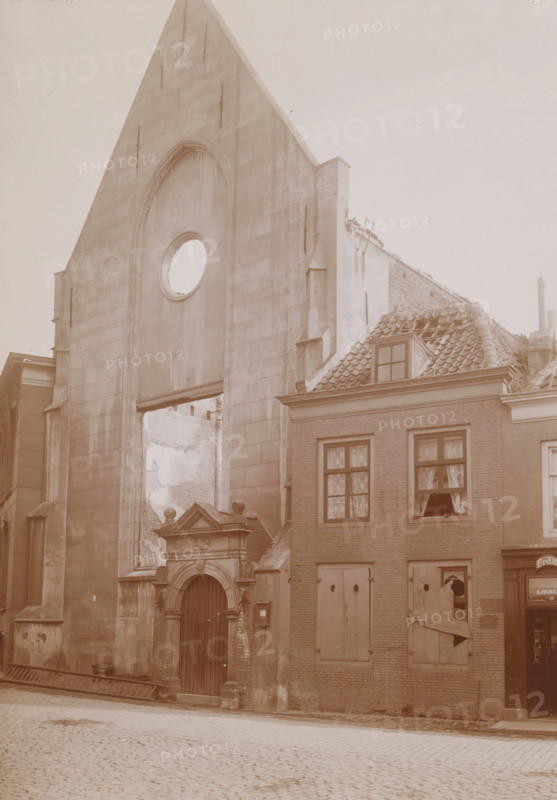 View of the Reformed Church in Vlissingen after the fire in 1911, photograph, annotation: 'NH church in Vlissingen after the fire of September 6. 1911', annotation: 'P. Louw, Ca. 1710', Flushing, 1911, photographer, Attributed to, paper, pasteboard, daylight collodion silver print, height 214 mm x width 152 mm, church (exterior), A tall stone façade with a pointed gable and a large circular opening sits next to a brick building with a tiled roof and a dormer window, the stone façade showing an arched recess and a decorated doorway with columns and a wooden door, while the adjacent brick building has shuttered and broken windows with curtains visible behind some panes and a boarded lower window, roof tiles appearing damaged and partially missing, rubble and exposed interior walls visible through gaps, and a cobblestone street in the foreground.