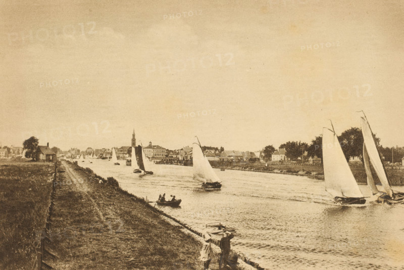 Sailing in North Holland, Sailing race on a waterway in North Holland, album page, photomechanical print, This print is part of an album., photographer: E. van Elfrinkhoff, North Holland, cliché maker: Nederlandsche Rotogravure Maatschappij, Leiden, in or before 1915, paper, rotogravure, height 161 mm x width 240 mm, sailing-ship, sailing-boat, river, A riverside scene in a muted sepia tone showing a stretch of grassy bank and a dirt path alongside a waterway with multiple sailboats of varying sizes on the water. Several boats have tall sails, some angled and some nearly vertical, and a small rowboat with figures is near the shore. Buildings and trees line the opposite bank, including a tower with a pointed top rising above nearby structures. Two people stand on the near bank looking toward the river, and scattered small boats are moored along the edge of the water.