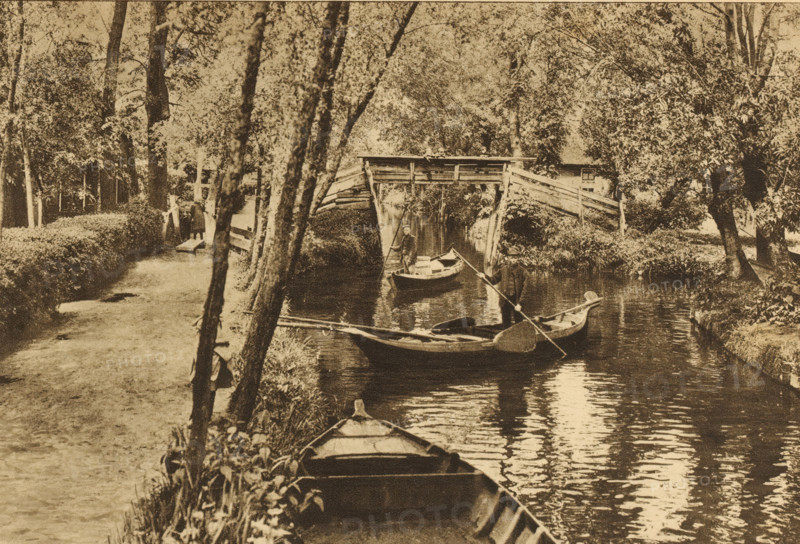 View of Giethoorn, Overijssel, Giethoorn, album page, photomechanical print, This print is part of an album., cliché maker: Nederlandsche Rotogravure Maatschappij, Leiden, in or before 1915, photographer, paper, rotogravure, height 161 mm x width 240 mm, prospect of village, silhouette of village, canals, waters (in city), gondola, A sepia-toned scene of a narrow river flanked by trees and vegetation with a wooden footpath running alongside one bank, several small rowboats on the water including one in the foreground tied to the bank, two men standing in separate boats holding long poles, a rustic wooden bridge arching over the river in the midground, dense foliage and tall trees on both sides, and reflections of the boats and trees visible in the water.