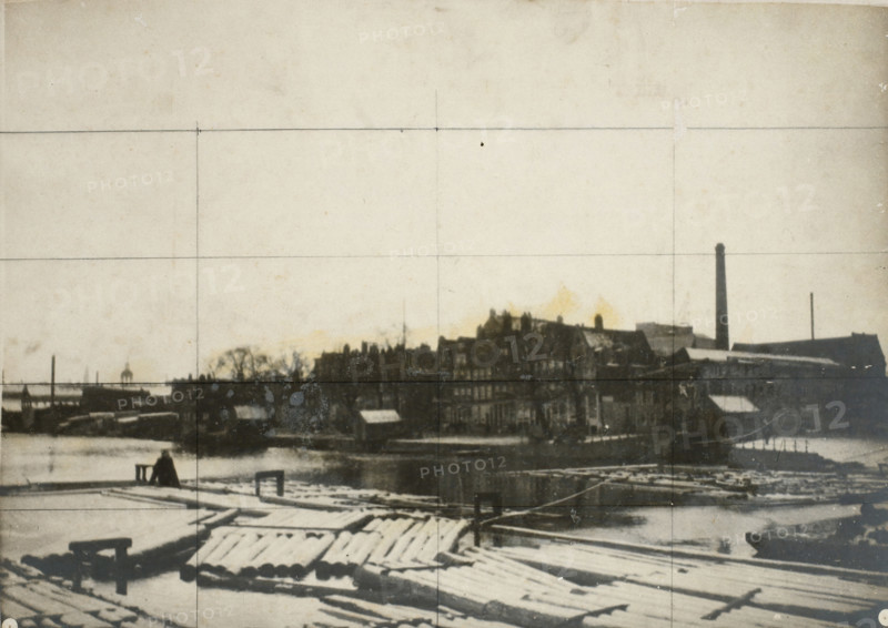 Westerdok under the snow in Amsterdam (former title), De Zandhoek in Amsterdam (former title), The Westerdok viewed towards the Zandhoek, photograph, The Westerdok and the Zandhoek under the snow in Amsterdam. Grid lines have been drawn on the photo in pencil., inscription: '84 x 158 1, 2', photographer: George Hendrik Breitner, Netherlands, 1902 - 1903, paper, pasteboard, gelatin silver print, developed gelatin silver print, height 361 mm x width 501 mm, height 380 mm x width 515 mm, harbour, Amsterdam, Westerdok, Zandhoek, A wide view of a waterfront scene with a wooden dock in the foreground showing planks and stacked logs partially covered in snow, a small bench or bollard near the water's edge, and a lone seated figure facing the water; across the water a dark silhouette of industrial and residential buildings lines the shore with rooftops and a tall chimney rising above them, a smaller slender smokestack further along, and leafless tree shapes interspersed among the structures; t...