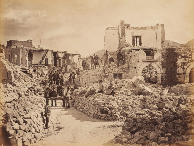 Destruction by an earthquake in southern Italy, photograph, annotation: 'f 30, -', inscription: 'Middle East 1890's', Italy (possibly), 1905 - 1908, photographer, Italy, pasteboard, paper, albumen print, albumendruk, height 188 mm x width 248 mm, height 238 mm x width 277 mm, ruin of a dwelling, house, castle, (carrying a person on a) litter, stretcher, consequences of an earthquake, Sicily, Calabria, A sepia-toned scene of ruined buildings and rubble filling a street, with crumbling stone walls, exposed interiors, and broken arches; several men stand or sit among the debris, some on higher piles of stone and others on the ground, a few appear to be carrying a stretcher-like object, and one man stands in the foreground holding a curved tool; scattered stones and broken masonry form makeshift low walls along the road, and remnants of doorways and window openings are visible in the damaged structures.