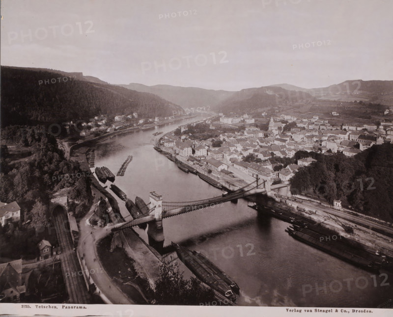 Panorama of the city of Decín (German: Tetschen-Bodenbach) on the Elbe, Czech Republic, photograph, Panorama of the city of Decín (German: Tetschen-Bodenbach) on the Elbe in the Czech Republic. Taken from a very high vantage point, placing the flow of the river and the location of the city in the center., inscription: 'Verlag von Stengel & Co., Dresden.', inscription: '3235. Tetschen, Panorama.', publisher: Stengel & Markert, Germany, c. 1890 - c. 1925, paper, photoaquatint, heliogravure, height 229 mm, width 284 mm, Severocesky Kraj, Elbe, Tetschen, A wide aerial view of a river winding through a town with a suspension bridge crossing the water, stone bridge towers at each end, and several long flat boats moored along the riverbanks. On one side of the river a dense forested hillside rises, dotted with small houses near the shore and a tunnel entrance visible at the base of the hill. On the opposite bank a compact cluster of buildings with pitched roofs and a church with a tower fill ...