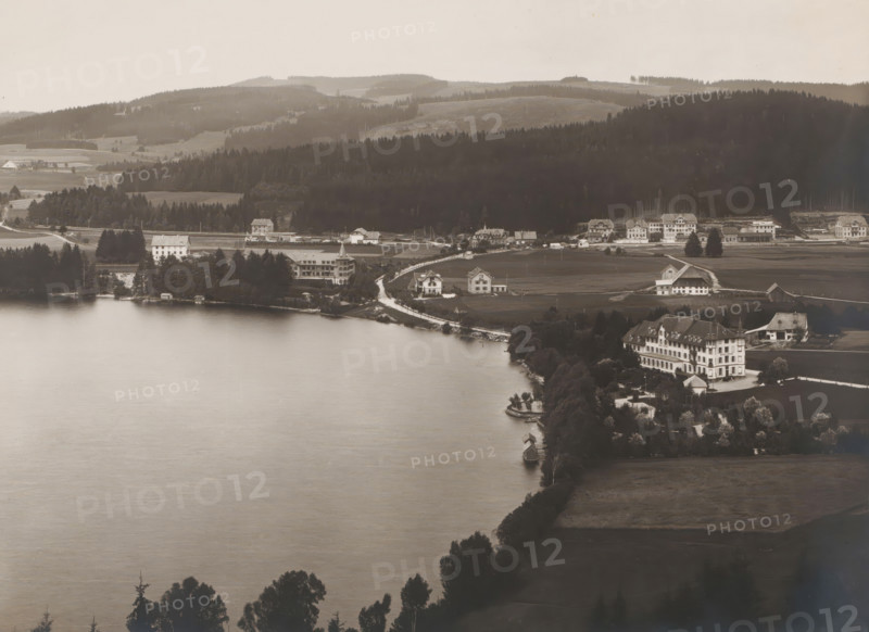 Titisee, Black Forest, Baden-Württemberg, Germany, photograph, The Black Forest lies on the Rhine and on the French border. On the northern shore of the lake, which covers 1.3 km², lies the spa town of the same name, nowadays part of the city of Titisee-Neustadt. Here we see Titisee from the South., number: '2313', inscription: 'Verlag der Neuen Photogr.Gesellschaft A.-G., Berlin-Stieglitz 1904', inscription: 'SCHWARZWALD. Titisee von Süden.', publisher: Photographische Gesellschaft Berlin, in or before 1904, paper, gelatin silver print, developing gelatin silver print, height 190 mm, width 242 mm, Titisee-Neustadt, A wide view of a lakeshore with calm water in the foreground and a shoreline dotted with trees, small docks, and boathouses, a curved road running along the shore past several standalone houses and larger multi-story buildings with pitched roofs, clusters of trees and manicured lawns around the buildings, open fields and farmhouses extending inland, and sloping forested hil...