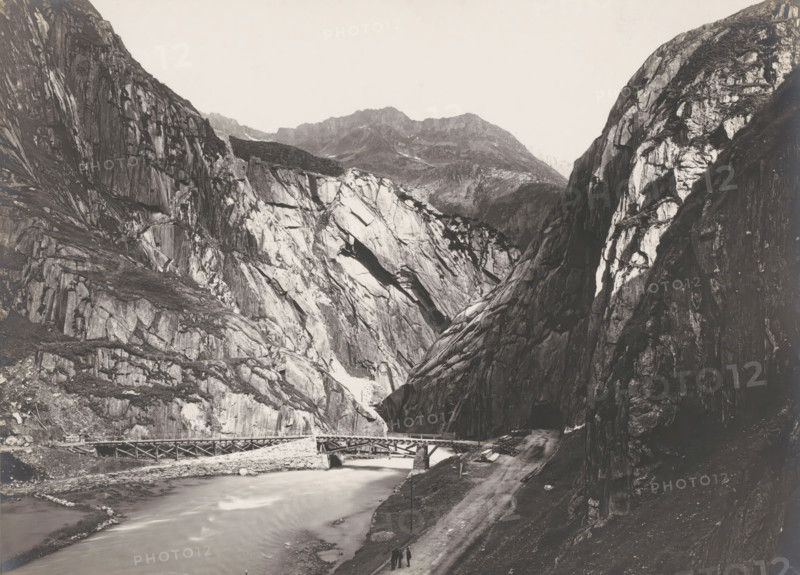 Tunnel 'Das Urnerloch' Schöllenen? and bridge over the Reuss, Gotthard Pass, Switzerland, photograph, To the right of the deep valley between steep rocks you can see the entrance to the 64-meter-long tunnel 'Urnerloch', built between 20. 09. 1707 and 15.08. 1708. A low bridge can be seen over the river Reuss., inscription: '2073. Das URNERLOCH.', inscription: 'Gebr. Wehrli, Kilchberg, Zurich.', photographer: Gebrüder Wehrli, c. 1900 - c. 1925, pasteboard, paper, gelatin silver print, developing gelatin silver print, height 161 mm, width 216 mm, A wide monochrome landscape showing steep rocky cliffs and craggy mountains with striated rock faces and lighter patches of exposed stone, a river flowing through the valley with a wooden bridge crossing it, a dirt road running alongside the river, and three small figures walking on the road near the foreground.