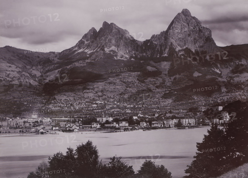 The place Brunnen and the two 'Myths' (mountain range), Switzerland, photograph, Brunnen is situated on Lake Lucerne in Central Switzerland. In the background, two striking mountain peaks - 'the two Myths' - can be seen., inscription: '1532. Brunnen & die Mythen', photographer: attributed to Gebrüder Wehrli, c. 1900 - c. 1920, Attributed to, paper, gelatin silver print, height 218 mm, width 165 mm, Brunnen, A wide grayscale landscape shows a large mountain range rising behind a tiered hillside dotted with trees and patches of lighter rock, with steep rocky peaks and ridges under a cloudy sky; below the slopes a built-up area with rows of multi-story buildings and smaller houses lines the shore of a calm body of water, where a small pier and a few boathouses project into the water; foreground trees frame the bottom edge of the image.