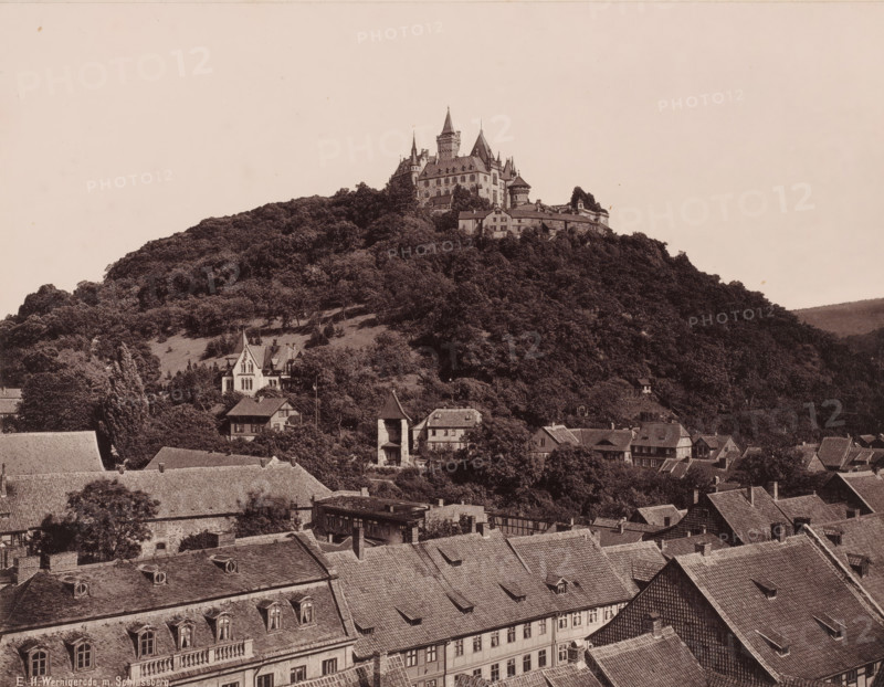 Wernigerode mit Schlossberg', Saxony-Anhalt, Germany, photograph, View of a castle located on a wooded mountain, with the roofs of the houses of Wernigerode in the foreground., inscription: 'E 11. Wernigerode m. Schlossberg.', c. 1885 - c. 1910, photographer, pasteboard, paper, albumen print, height 212 mm x width 274 mm, height 356 mm x width 455 mm, Wernigerode, A hill densely covered in trees rises above a town, topped by a large stone castle complex with multiple steep roofs, towers, turrets, and crenellated walls, while below the hill a cluster of houses and buildings with pitched roofs, dormer windows, and chimneys sit among trees and small clearings.