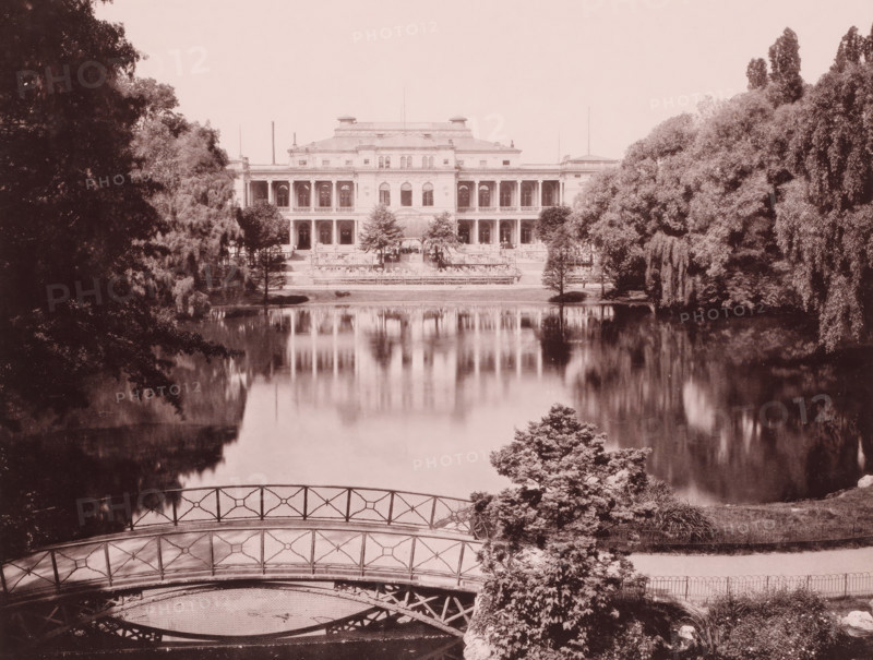 House in a park by a large pond, Frankfurt am Main ?, Germany, photograph, Germany., c. 1885 - c. 1895, photographer, pasteboard, paper, albumen print, height 202 mm x width 268 mm, height 508 mm x width 330 mm, A wide, symmetrical building with two stories of columned balconies and a central entrance sits at the far edge of a calm reflective body of water, its facade mirrored in the lake; flanking the building are dense clusters of tall trees and shrubs, with large trees framing the scene on both sides. In the foreground a decorative metal footbridge with geometric railing arches over a small inlet, and near the bridge a leafy bush and other low plants grow beside a gently sloping bank. The overall image has a sepia tone and soft, slightly faded contrast, with reflections of the building and surrounding trees visible on the water surface.