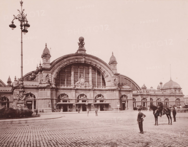 Equestrian statue on a pedestal of Emperor Wilhelm I, Frankfurt-am-Main, Germany, photograph, Germany. Sculptor was Clemens Buscher (1855–1916), who created the statue in 1896. It was destroyed and melted down during WWII., caption: 'Station Frankfurt', c. 1875 - c. 1900, photographer, paper, pasteboard, collotype, photogravure on photographic paper on cardboard, height 274 mm, width 202 mm x width 490 mm, height 330 mm, A large stone building with an ornate arched central facade and tall arched windows topped by sculpted figures and decorative carvings spans the scene, flanked by domed towers and smaller ornamental turrets; a tall ornate streetlamp rises to the left foreground, and a cobbled plaza stretches across the front. Several people stand near the building's entrances and in the plaza, and a man in a suit stands nearer to the camera with a horse and a handler slightly behind him to the right. The sky is empty above the building, and architectural details such as columns, statue...