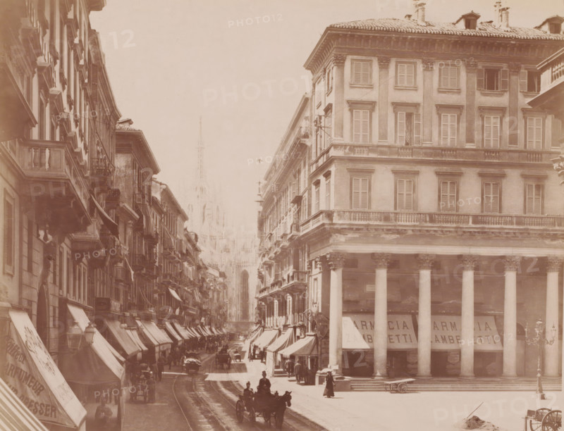 Corso Vittorio Emanuele, Milan, Italy, photograph, View of the street Corso Vittorio Emanuele with shop fronts, horse and carriages and pedestrians, Italy., caption: '6830a MILANO. Corso Vittorio Emanuele.', caption: '(EDIZIONI BROGI)', photographer: Edizioni Brogi, Florence, publisher: Giacomo Brogi, c. 1860 - c. 1890, Tuscany, pasteboard, paper, albumen print, height 197 mm x width 249 mm, height 492 mm x width 330 mm, Lombardy, A sepia-toned street scene showing tall multi-story buildings lining a narrow avenue, with rows of shuttered windows and ornate balconies, awnings extending over the sidewalks, and a colonnaded building with large columns on the right; a distant Gothic-style cathedral spire is visible through a light haze at the end of the street, while horse-drawn carriages and pedestrians occupy the roadway and sidewalks, tram rails running down the center, and street lamps and signage projecting from building facades.