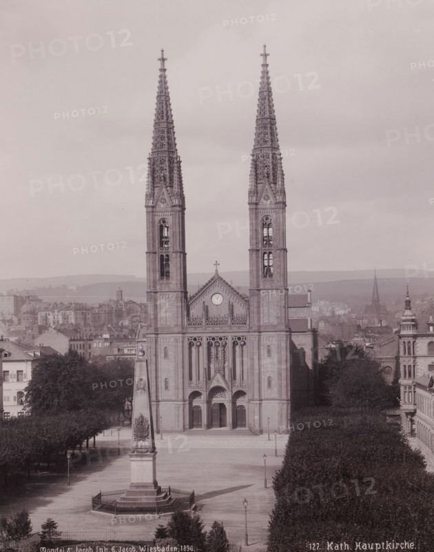 Katholische Hauptkirche', Wiesbaden, Germany, photograph, Frontal view of the church and the empty square in front of it from a high point of view., inscription: '127. Kat. Hauptkirche.', inscription: 'Mondel & Jacob Inh. G. Jacob, Hof-Phot. Wiesbaden 1896.', photographer: Mondel & Jacob, photographer: Mondel, photographer: Julius Jacob, 1896, Mondel & Jacob, Mondel, Julius Jacob, photographer, Hessen, pasteboard, paper, gelatin silver print, developing gelatin silver print, height 282 mm, width 225 mm x width 330 mm, height 508 mm, A large stone church façade with two tall ornate spires dominates the center of the image, each spire featuring intricate Gothic tracery and pointed finials, and the central section between them showing a triangular gable with a circular clock and a cross at its peak; below the gable are tall arched doorways and detailed sculptural relief above the main entrance. In front of the church is an open square with a tall obelisk monument on a stepped base enclose...